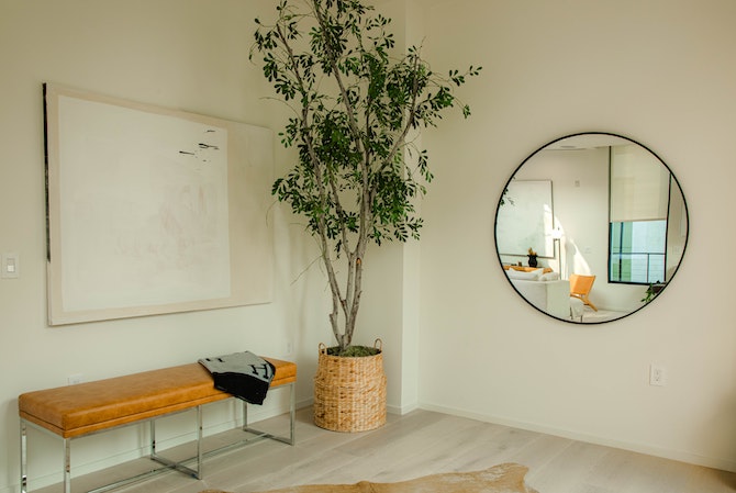 Okruglo ogledalo na zidu hodnika pored koga se nalazi visoka biljka u saksiji pored kožne klupice